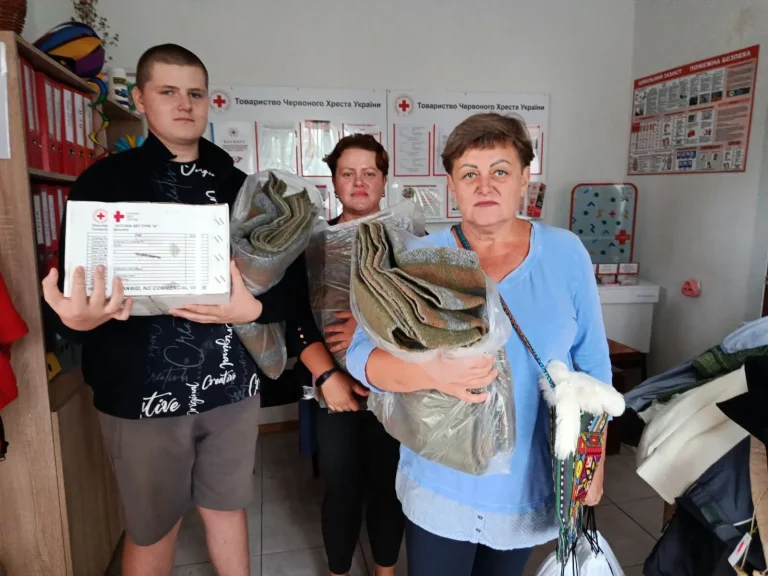 Троє людей у приміщенні Червоного Хреста тримають гуманітарну коробку та ковдри