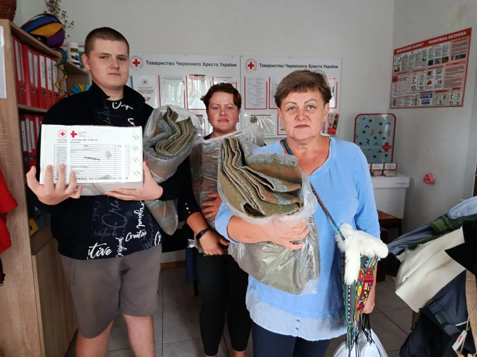 Троє людей у приміщенні Червоного Хреста тримають гуманітарну коробку та ковдри